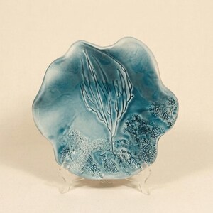 Peut inclure: Une assiette en céramique bleu-vert faite à la main, de forme organique unique. L'assiette présente un motif central en relief ressemblant à un arbre ou une plante stylisée, avec des détails texturés et une finition brillante. L'assiette est présentée sur un support transparent.