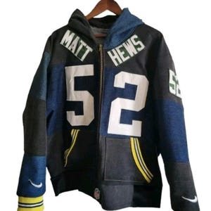 May include: A zip-up hoodie with a patchwork design in shades of blue, gray, and black. The hoodie features the name "MATT HEWS" and the number "52" in white. Yellow stripes accent the pockets and cuffs, with a Nike logo on the sleeve.