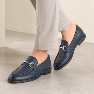 May include: A pair of navy blue leather loafers featuring a silver-toned metal horsebit detail across the vamp. The shoes have a rounded toe, a low stacked heel, and a smooth finish, paired with light gray trousers that fall above the ankles.