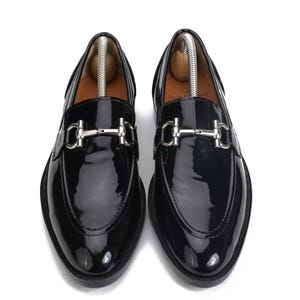 May include: A pair of black patent leather loafers featuring a silver-toned metal horsebit detail across the vamp. The shoes have a rounded toe, a smooth glossy finish, and include wooden shoe trees inserted inside to maintain their shape.