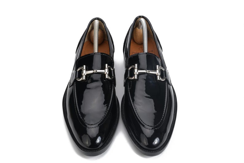 May include: A pair of black patent leather loafers featuring a silver-toned metal horsebit detail across the vamp. The shoes have a rounded toe, a smooth glossy finish, and include wooden shoe trees inserted inside to maintain their shape.