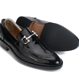 May include: A pair of black patent leather loafers featuring a silver-toned metal horsebit detail across the vamp. One shoe is positioned upright with a metal shoe tree inside, while the other is placed on its side to display the black textured sole.