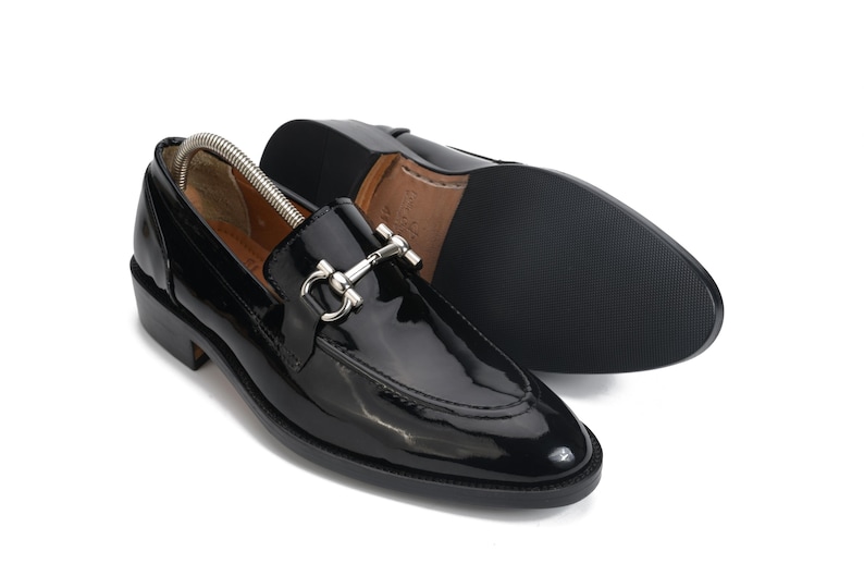 May include: A pair of black patent leather loafers featuring a silver-toned metal horsebit detail across the vamp. One shoe is positioned upright with a metal shoe tree inside, while the other is placed on its side to display the black textured sole.