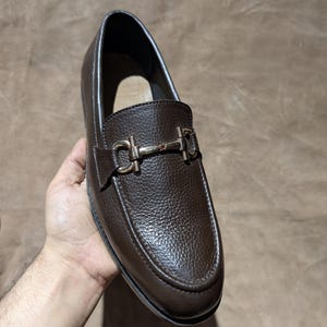 May include: A dark brown leather loafer featuring a textured pebble grain finish and a polished silver-tone metal horsebit detail across the vamp. The shoe has a rounded toe, a structured silhouette, and a smooth interior lining.