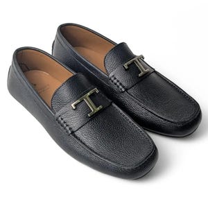 May include: A pair of black leather loafers with a pebbled texture, featuring a silver-toned metal T-shaped ornament across the vamp. The interior of the shoes is lined with a light tan material, and the design includes a rounded toe and stitching.