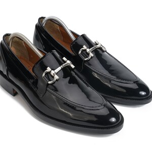 May include: A pair of black patent leather loafers featuring a polished silver-tone metal horsebit detail across the vamp. The shoes have a rounded toe, a low stacked heel, and a black sole, with silver metal shoe trees inserted inside each shoe.