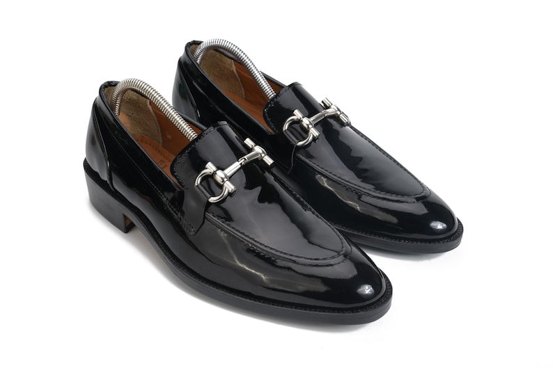 May include: A pair of black patent leather loafers featuring a polished silver-tone metal horsebit detail across the vamp. The shoes have a rounded toe, a low stacked heel, and a black sole, with silver metal shoe trees inserted inside each shoe.