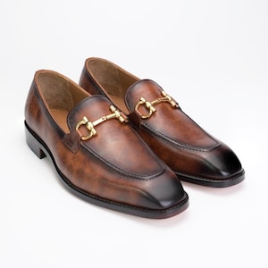 May include: A pair of brown leather loafers featuring a burnished finish with darker tones at the toe and heel. Each shoe is accented with a polished gold-tone metal horsebit detail across the vamp, set on a low heel with a smooth sole.
