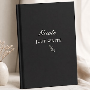 May include: A black hardcover notebook featuring the name Nicole in a white script font, followed by the text JUST WRITE in a white sans-serif font, and a small white line drawing of a leafy branch centered below the text on the front cover.