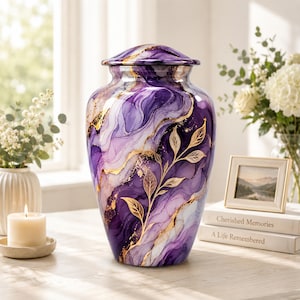 May include: A decorative urn featuring a swirling marble pattern in shades of deep purple, lavender, and white, accented with metallic gold veins and a delicate gold leaf branch design. The vessel has a rounded body and a matching removable lid.