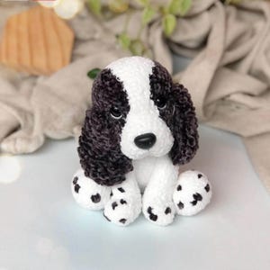 Könnte beinhalten: Ein handgefertigter Plüschhund in Schwarz und Weiß. Der Hund hat ein weißes Gesicht und weiße Pfoten mit schwarzen Flecken. Die Ohren und der Kopf sind schwarz. Der Hund sitzt auf einer weißen Oberfläche.