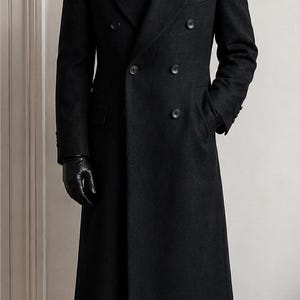 Puede incluir: Un abrigo cruzado de lana gris carbón con solapas de pico anchas, seis botones frontales y dos bolsillos laterales. El abrigo se lleva sobre una camisa blanca de cuello, una corbata verde oliva, pantalones negros, guantes de cuero negros y zapatos.