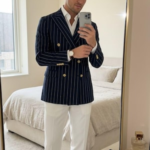 Pode incluir: Um blazer azul-marinho de abotoamento duplo com riscas brancas e botões dourados, combinado com uma camisa branca de colarinho, calças brancas e mocassins de couro preto com borlas. Um relógio de pulso dourado é usado no pulso esquerdo.