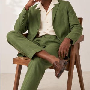 Op de afbeelding: Een olijfgroen pak bestaande uit een single-breasted blazer met inkeping revers en een bijpassende broek, gecombineerd met een crèmekleurig overhemd met korte mouwen en een camp-kraag en bruine leren loafers.