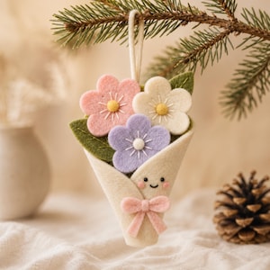 May include: A felt hanging ornament shaped like a bouquet of three flowers in pink, cream, and lavender. The flowers have yellow or white centres and green felt leaves. The bouquet is wrapped in cream felt with a small smiling face and a pink bow.