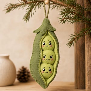 May include: A felt hanging ornament shaped like a green pea pod containing three individual peas. The top and bottom peas have sad expressions with downturned mouths, while the middle pea has an angry expression with furrowed eyebrows.