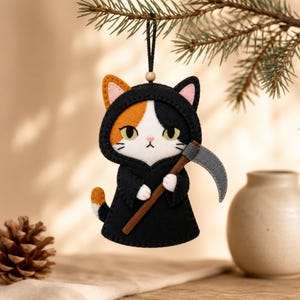 May include: A felt hanging ornament shaped like a calico cat dressed in a black hooded robe. The cat has white, orange, and black fur, yellow eyes, and holds a grey scythe with a brown handle. It hangs from a black string with a small wooden bead.