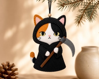 Könnte beinhalten: Ein hängendes Filzornament in Form einer dreifarbigen Katze, die eine schwarze Kapuzenrobe trägt. Die Katze hat weißes, orangefarbenes und schwarzes Fell, gelbe Augen und hält eine graue Sense mit braunem Stiel. Sie hängt an einer schwarzen Schnur.