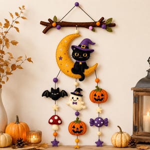 Peut inclure: Une suspension murale en feutrine avec une branche brune ornée de pompons violets et orange. Un croissant de lune jaune avec un chat noir portant un chapeau de sorcière violet est suspendu, avec des formes en feutrine de chauve-souris, fantôme, citrouilles et bonbons.