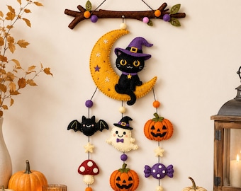 May include: A felt wall hanging featuring a brown branch at the top with purple and orange pom-poms. A yellow crescent moon with a black cat in a purple witch hat hangs below, followed by felt shapes including a bat, ghost, pumpkins, and candy.