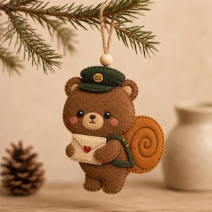 Puede incluir: Un adorno de fieltro con forma de oso marrón que lleva una gorra de cartero verde oscuro con un icono de sobre amarillo. El oso sostiene un sobre color crema con un pequeño corazón rojo y lleva un bolso verde oscuro. Cuelga de un cordel beige.