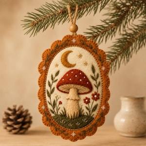 May include: An oval felt ornament featuring an embroidered red mushroom with white spots, a yellow crescent moon, and small stars. The design is framed by a scalloped burnt orange border with green foliage and small red flowers, hanging from a jute string.