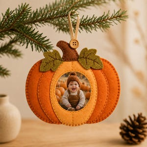 Puede incluir: Un adorno fotográfico de fieltro con forma de calabaza que presenta un recorte circular central con la foto de un bebé sonriente con gorro y peto marrones. El adorno tiene un tallo marrón, dos hojas verdes y un lazo beige con un botón.