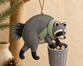 May include: A felt raccoon ornament featuring a grey body, black tail, and a light green scarf. The raccoon is reaching into a small grey felt trash can. The ornament is suspended by a brown string with a small wooden bead at the top.