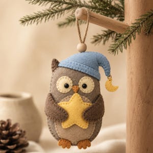 May include: A felt owl ornament in shades of brown, wearing a light blue nightcap with a small yellow crescent moon charm attached. The owl has cream-colored eye patches, black eyes, and holds a yellow star in its wings. A wooden bead and twine loop are attached to the top for hanging.