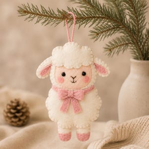 May include: A felt ornament shaped like a lamb with cream-coloured wool, pink inner ears, pink cheeks, and a pink bow around its neck. The lamb has black embroidered eyes and a nose, and it hangs from a pink string loop for display purposes.