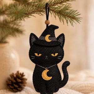 Puede incluir: Un adorno de fieltro negro con forma de gato que lleva un sombrero de bruja puntiagudo con un detalle de media luna amarilla. El gato tiene ojos amarillos, bigotes bordados y un dije de media luna amarilla colgado de un cordón oscuro.