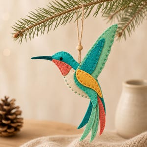 Può includere: Un ornamento in feltro a forma di colibrì con strati di tessuto color verde acqua, verde chiaro, giallo e corallo, rifiniti con cuciture decorative a vista. L'uccello ha un piccolo occhio a perla nera e pende da un filo con perla.