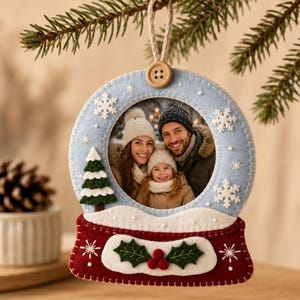 May include: A felt snow globe ornament featuring a light blue frame with white embroidered snowflakes and a wooden button at the top. The base is dark red with white felt trim, green holly leaves, and red berries. It holds a photo of three people.