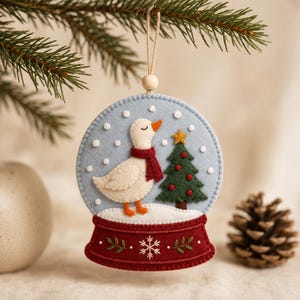 Pode incluir: Um ornamento de feltro em forma de globo de neve com um ganso branco com bico e patas cor de laranja, usando um cachecol vermelho. Ao lado do ganso está uma árvore de Natal verde com enfeites vermelhos e uma estrela amarela na base.
