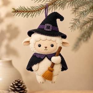May include: A felt ornament shaped like a cream-colored sheep wearing a black witch hat with a purple band and a matching black cape with a purple collar. The sheep holds a small brown and orange broomstick, and a purple string is attached to the top.