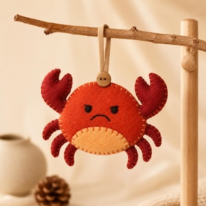 May include: A felt crab ornament in vibrant red and orange, featuring a grumpy embroidered face with a downturned mouth and furrowed brows. The crab has two raised claws and six legs, attached to a beige fabric loop with a small wooden button.