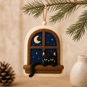 Op de afbeelding: Een vilten hangornament in de vorm van een boogvenster met een bruin frame. Een zwarte kat zit op de bruine vensterbank tegen een donkerblauwe nachtelijke hemel met een gele maansikkel en kleine witte sterretjes. Met een beige touwtje.