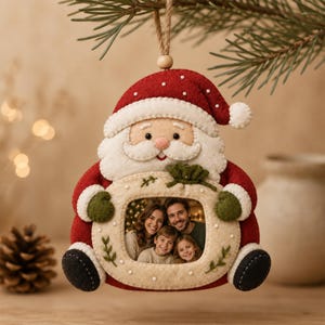 Könnte beinhalten: Ein Weihnachtsanhänger aus Filz in Form eines Weihnachtsmanns mit einem ovalen Fotorahmen in der Mitte. Die Figur trägt eine rote Mütze und einen Mantel mit weißem Besatz, grüne Fäustlinge und dunkelgraue Stiefel. Eine grüne Schleife ziert den Rahmen.