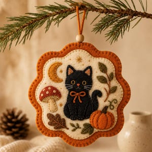 Könnte beinhalten: Ein wellenförmiger Filzanhänger mit einem gestickten schwarzen Katzenmotiv mit Schleife, einer Mondsichel, einem roten Pilz, einem orangefarbenen Kürbis und Herbstblättern. Die cremefarbene Mitte ist von einem rostorangen Rand umgeben.