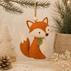 May include: A felt fox ornament in burnt orange and cream, featuring a small green leaf accent around the neck. The fox has stitched details, a dark brown nose, and is suspended by a jute twine loop with a single light-coloured wooden bead.
