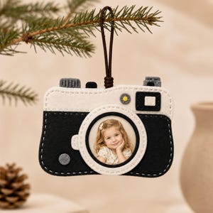 May include: A felt camera-shaped ornament featuring a black and cream colour scheme with white stitching. The centre lens area displays a circular photograph of a young child. A brown cord is attached to the top for hanging purposes.