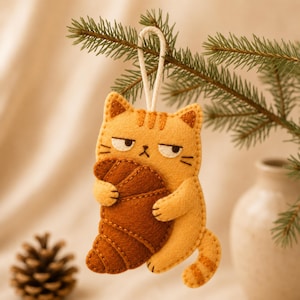 May include: A felt hanging ornament shaped like a light orange tabby cat with a grumpy expression, holding a dark brown croissant. The cat has stitched details, a curved tail, and a white cord loop at the top for hanging from a tree branch.