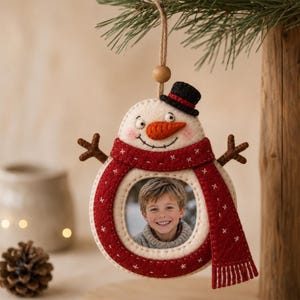 May include: A felt snowman-shaped photo frame ornament featuring a cream-colored body, a bright orange carrot nose, a small black top hat, and a long red scarf with white stitched details. The center holds a circular photo of a young child.