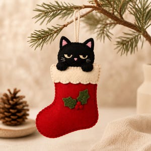 May include: A felt Christmas ornament shaped like a red stocking with a white cuff, featuring a black cat with pink inner ears and a pink nose peeking out from the top. The stocking is decorated with green holly leaves and three small red berries.