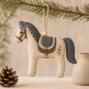 May include: A felt horse ornament in cream with a dark grey mane, tail, and saddle. The horse features a brown bridle, a small wooden bead on the hanging loop, and a delicate green leaf embroidery on its flank. The edges are finished with visible stitching.