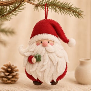 Pode incluir: Um enfeite de feltro do Pai Natal com um gorro vermelho de pompom branco, uma barba branca espessa e uma pequena caixa de presente verde com um laço branco na mão. O enfeite está suspenso por um fio vermelho e tem pés castanhos na base.