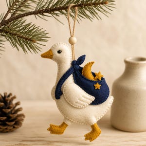 Op de afbeelding: Een vilten ornament in de vorm van een witte gans met een gele snavel en gele poten. De gans draagt een donkerblauwe tas met daarin een gele halve maan en twee kleine gele sterren. Aan de bovenkant zit een beige touwtje met een houten kraal.