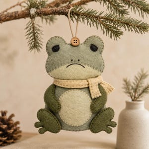 Può includere: Un ornamento in feltro a forma di rana nei toni del verde salvia e oliva, con pancia e sciarpa color crema. La rana presenta occhi neri ricamati, una semplice linea per la bocca ed è appesa a un cordino beige con un piccolo bottone in legno.
