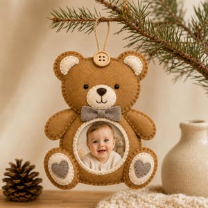May include: A brown felt teddy bear ornament featuring a circular photo frame in the center displaying a baby's portrait. The bear has cream-colored ear accents, a grey bow tie, a wooden button at the top, and grey heart patches on its feet.