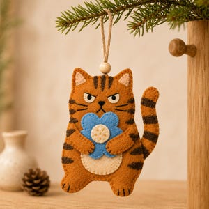 May include: A felt cat ornament featuring a grumpy expression, orange fur with dark brown stripes, and a cream-coloured belly. The cat holds a light blue flower with a cream centre. It hangs from a jute string with a small wooden bead at the top.